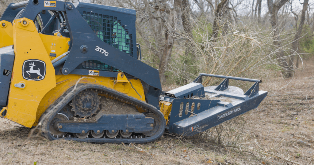 A tracked skid steer loader clearing brush in a wooded area.