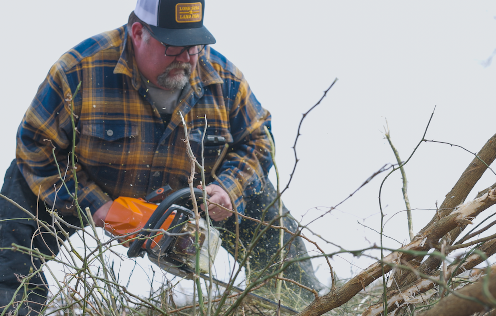 Man in plaid shirt using chainsaw to cut tree branches outdoors.